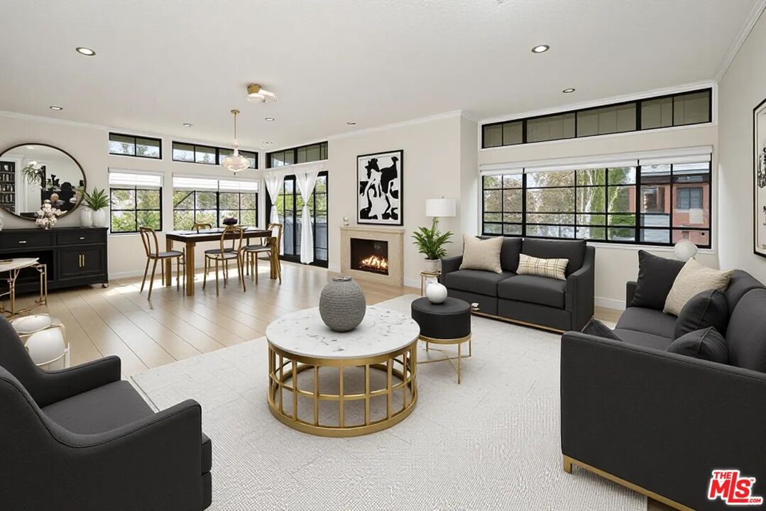 Sun-filled remodeled living and dining room at 200 N Swall Drive #553, with a gas fireplace, gold chandelier, factory-style steel windows and treetop view through the terrace doors.
